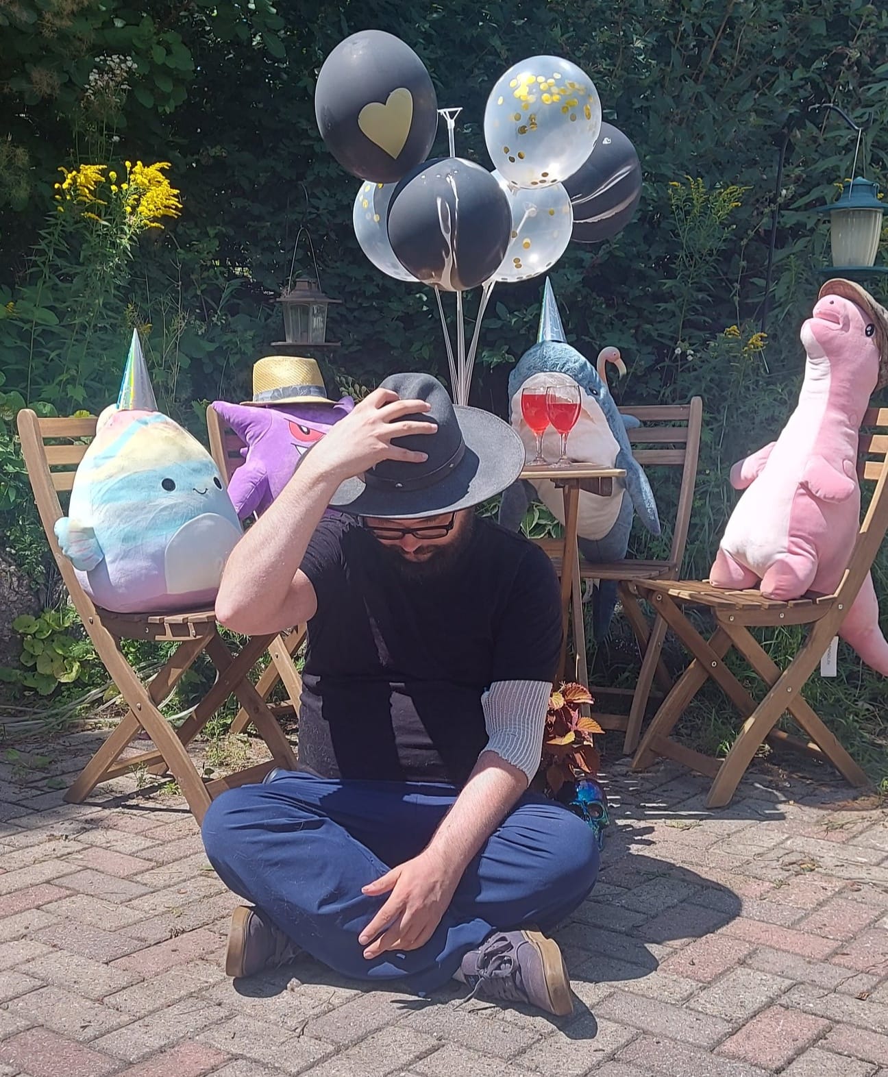 A man with facial hair and a felt sun hat sits on the ground in front of a patio set hosting a stuffed animal patio party, Photo 2